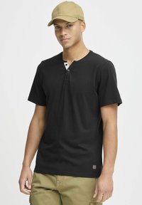Černé krátké rukávy henley tričko s knoflíkovým potiskem a kontrastními světlými knoflíky. Model nese čepici a cargo kalhoty, stojí na bílém pozadí.