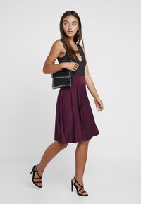 Fato preto justo com decote rendado, combinado com uma saia roxa fluida até aos joelhos. Acessorizado com uma bolsa preta de contas e sapatos de salto pretos.