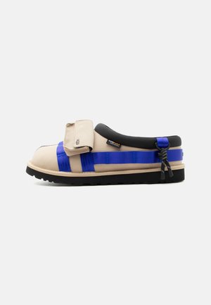 Zapato beige sin cordones con correas azules, cuello acolchado negro, solapa frontal con el logo de UGG y suela de goma negra sobre fondo blanco.
