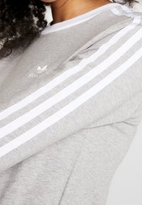 Grå sweatshirt med tre vita horisontella ränder längs ärmarna och en liten logotyp på bröstet. Mjuk bomullsblandning.