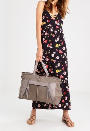 Žena v květovaných maxi šatech s ramínky drží velkou taupe semišovou tašku s třásněmi, má na sobě hnědé sandály a stojí na jednobarevném pozadí.