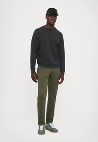 Mann som står og har på seg en svart caps, mørkegrå sweatshirt, olivenfargede bukser og grønne og hvite joggesko mot en enkel lys bakgrunn.