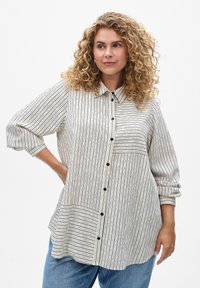 Chemise ample à manches longues en blanc cassé avec des rayures verticales noires. Comprend un devant à boutons et une poche latérale. Texture de tissu douce.