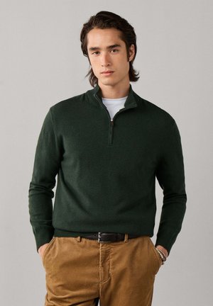 Joven con cabello oscuro de longitud media que lleva un jersey verde oscuro con cremallera sobre una camisa blanca y pantalones marrones con las manos en los bolsillos.