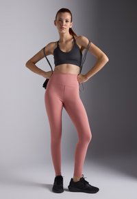 Svart sports-bh med justerbare stropper, kombinert med høytlivede, glatte, rosa tights. Modellen står med hendene på hoftene og har på seg svarte joggesko.
