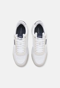 Hvite sneakers med beige detaljer, teksturert skinn og tekstil overdel, rund tå og flate snører. Har logo på tungen og sålen.