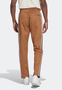 Pantalones marrones ajustados en la parte inferior con cintura elástica, que cuentan con franjas laterales moradas y un bolsillo trasero con cremallera. El material parece suave y ligero.