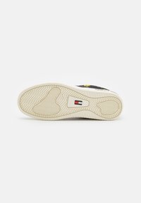 Suela de una zapatilla beige con patrones de tracción detallados y un pequeño logo rectangular en rojo, blanco y azul en el centro.