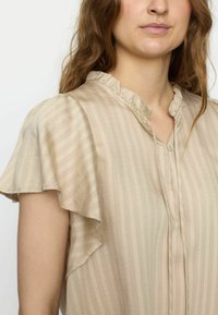 Beige blus med volangkrage och korta ärmar. Vertikala ränder och knäppning fram. Mjuk, texturerad tyg med en lätt och luftig känsla.