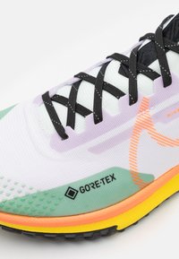 Nahaufnahme eines weißen, grünen und orangefarbenen Sneakers mit schwarzen Schnürsenkeln und Gore-Tex-Label, das atmungsaktives Mesh und strukturierte Sohlenmerkmale hervorhebt.