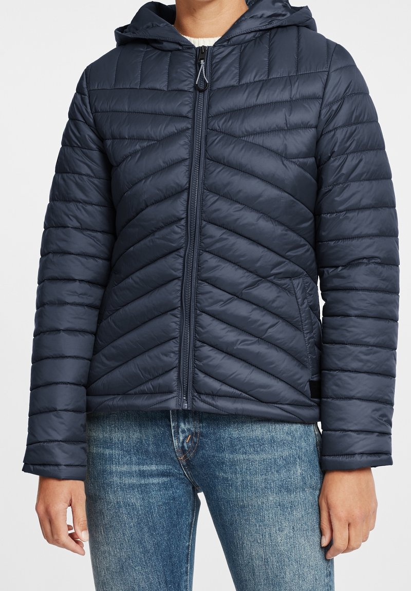 Veste matelassée à capuche bleu marine zippée, portée avec un jean bleu, montrant le torse et les bras d'une personne debout.