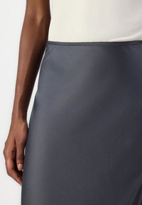 Osoba ubrana w ciemnoszary satynowy spódnicę i białą bluzkę, z lewą ręką swobodnie opartą na boku, na jednolitym tle.