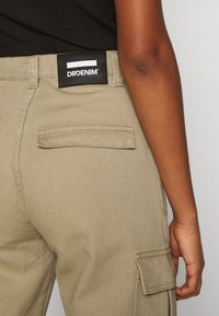 Kárované cargo kalhoty v béžové barvě s texturovaným povrchem, které mají černou značkovou nášivku na pase a zadní kapsu s klopou.