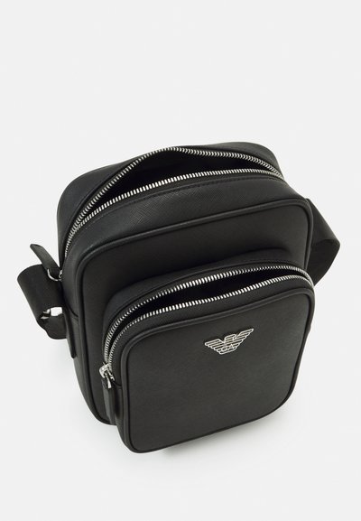Svart crossbody-väska i strukturerat material, med silverfärgade dragkedjor, två fack och en logotypdetalj på framsidan. Kompakt design.
