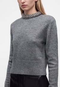 Femme portant un pull en maille gris avec un col orné de bijoux et un pantalon en cuir noir, visible du milieu du torse jusqu'au menton.