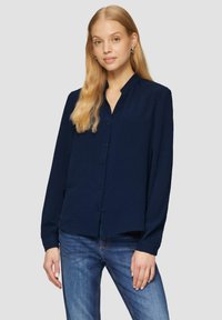 Dunkelblaues langärmliges Hemd mit strukturiertem Finish, Knopfleiste und kragenlosem Ausschnitt. Kombiniert mit hellblauen Jeans.