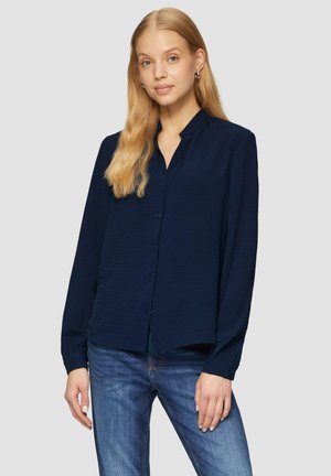 Marineblauwe longsleeve blouse met een gestructureerde afwerking, knoopsluiting aan de voorkant en een kraag. Gecombineerd met lichtblauwe jeans.