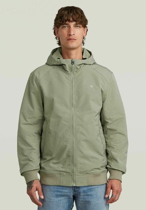 Jonge man met krullend haar, gekleed in een lichtgroen gewatteerd jack met capuchon en blauwe jeans, staand tegen een effen lichte achtergrond.