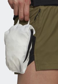 Main tenant une petite pochette blanche zippée avec une poignée en boucle, tenue à côté d'un short vert olive avec une poche zippée noire.