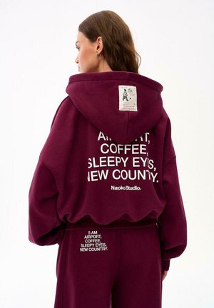 Kobieta o brązowych włosach w bordowej bluzie z kapturem i spodniach z białym napisem "5 AM Airport, Coffee, Sleepy Eyes, New Country" na plecach.