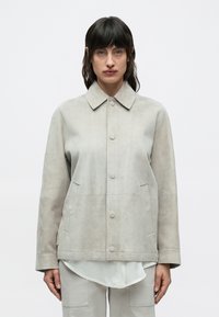 Junge Person mit dunklen Haaren, die eine hellgraue Wildlederjacke über einem weißen Hemd und passenden hellgrauen Hosen vor einem einfarbigen Hintergrund trägt.