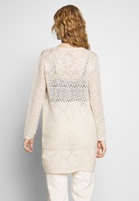 Langer, gestrickter Cardigan in Hellbeige mit geometrischem Muster. Verfügt über ein offenes Strickmuster, lange Ärmel und einen leicht geschwungenen Saum.