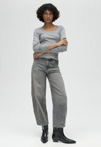 Donna con capelli ricci che indossa una maglietta grigia a maniche lunghe, jeans grigi sbiaditi a gamba larga e stivaletti neri, in piedi con le braccia incrociate.