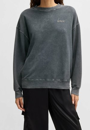 Frau trägt ein gewaschenes graues oversized Sweatshirt mit „boss“ auf der Brust bestickt und lockere schwarze Hosen.