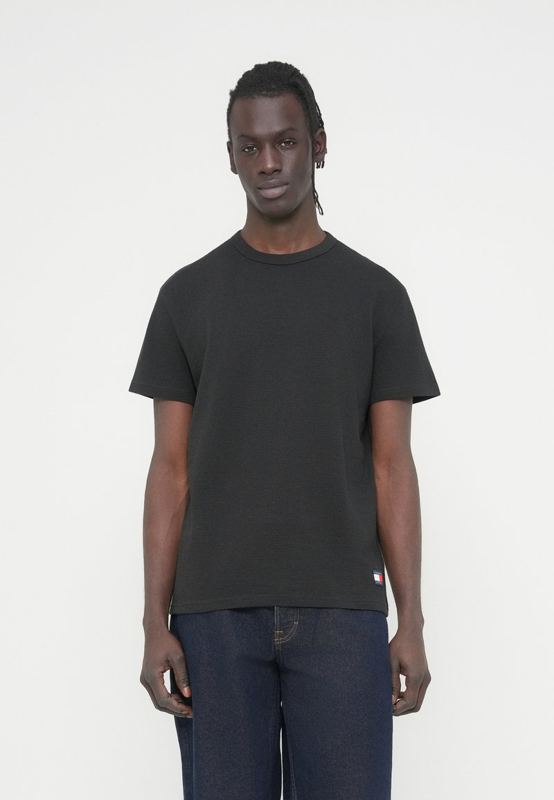 Μαύρο μπλουζάκι με στρογγυλή λαιμόκοψη σε υφασμάτινη υφή, κοντά μανίκια, με μικρή ετικέτα λογότυπου στο κάτω μέρος της ραφής. Συνδυασμένο με μπλε τζιν παντελόνια.