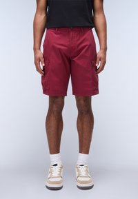 Pantaloni cargo bordeaux con due tasche laterali, realizzati in tessuto liscio. Abbinati a sneakers bianche e beige e calzini bianchi alla caviglia.