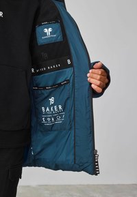 Veste matelassée bleue mettant en avant une poche technologique, des étiquettes de logo intérieures et un tissu texturé. Elle dispose d'une fermeture éclair et d'un tirette de zip marqué.