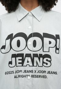 Πουκάμισο με ρίγες σε γκρι και λευκό, με μεγάλο μαύρο γραφικό "JOOP! JEANS" στο μπροστινό μέρος και λευκά κουμπιά κατά μήκος του λάστιχου.