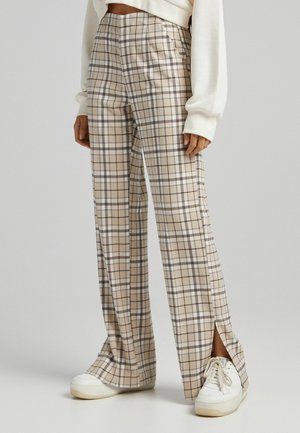 Beige geruite broek met een hoge taille, met zijsplitten bij de enkels, gemaakt van een soepele stof. Draag een paar witte sneakers.