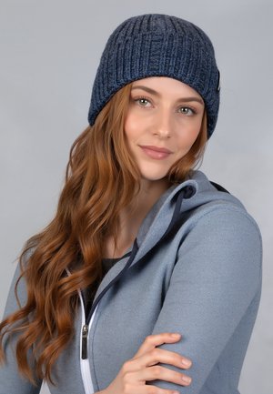 Gebreide marineblauwe beanie met ribbelstructuur; zacht materiaal; heeft een klein logo-label aan de zijkant. Gekleed met een grijze hoodie met rits.