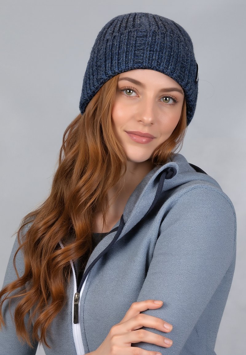 Gebreide marineblauwe beanie met ribbelstructuur; zacht materiaal; heeft een klein logo-label aan de zijkant. Gekleed met een grijze hoodie met rits.