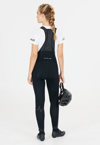Schwarze Fahrradhosen mit Netzträgern, versehen mit einem Logo und einem Herzschlagdesign an den Beinen. Trägt einen schwarzen Helm, der in einer Hand gehalten wird.