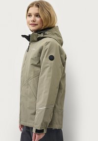 Beige waterdichte jas met een capuchon, ritssluiting, elastische manchetten en een zwart rond logo op de mouw. Kenmerkt zich door een gestructureerde afwerking.