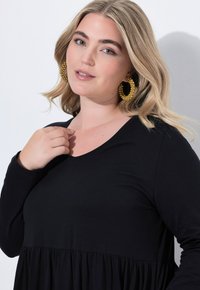 Haut noir à manches longues avec un décolleté en V, présentant un ourlet élastiqué et évasé. Le modèle porte de grandes boucles d'oreilles rondes en perles jaunes.
