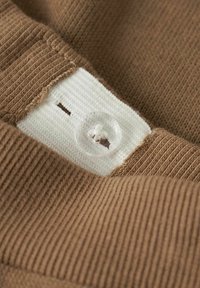 Ruskea neulottu kangas, jossa resorikuvio, valkoinen paikka ja kirkas nappi. Lähikuva korostaa tekstuuria ja napin yksityiskohtia.