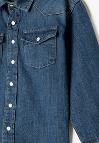 Blå denimskjorta med knapprad fram, två bröstfickor och klassisk krage. Texturerad tyg med kontrasterande sömnadsdetaljer.