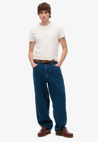 Camiseta blanca de manga corta, jeans de mezclilla azul de corte holgado, cinturón marrón, zapatos de barco marrones. Modelo de pie con las manos en los bolsillos sobre un fondo liso.