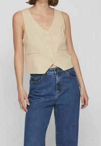 Béžová vesta bez rukávů s výstřihem do V, pěti knoflíky a dvěma předními kapsami, kombinovaná s modrými džínami s vysokým pasem.