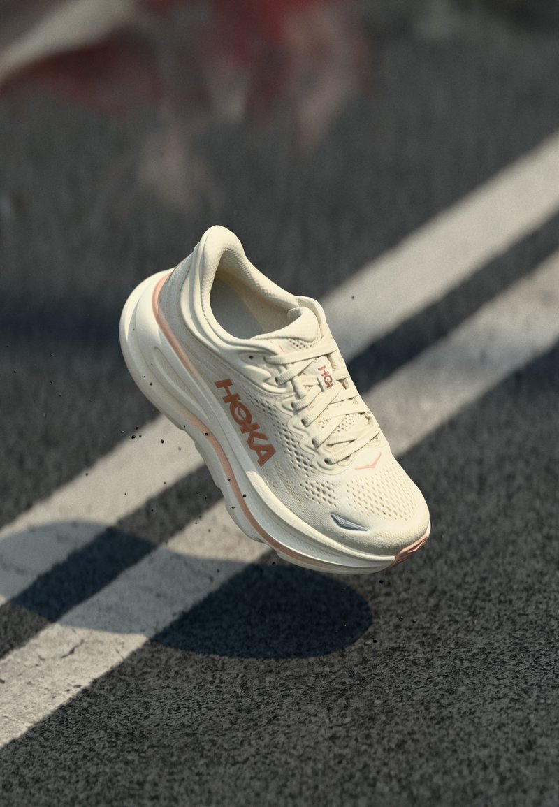 Lys beige Hoka løpesko med ferskenfarget logo, svevende over asfalt med hvit linje i dagslys.
