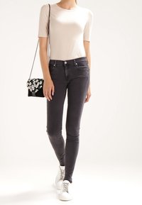 Mujer con blusa beige de manga corta, jeans ajustados oscuros, zapatillas blancas y un bolso cruzado negro con flores y cadena.