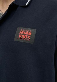Navyblaues Polohemd mit strukturiertem Stoff, versehen mit einem roten "JACK & JONES"-Logo auf einem rechteckigen Patch und einem weißen Streifen entlang des Kragens.
