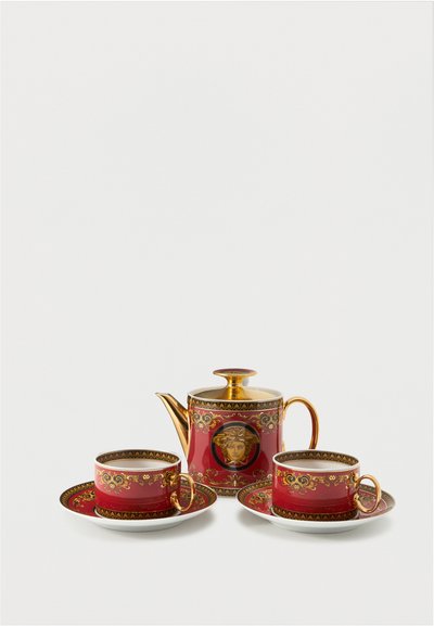 Théière ornée rouge et or avec emblème de Méduse entre deux tasses assorties sur des soucoupes, sur fond blanc.