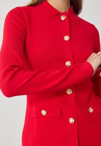 Primer plano de una chaqueta roja de manga larga con botones dorados ornamentales y bolsillos de solapa en la parte delantera.