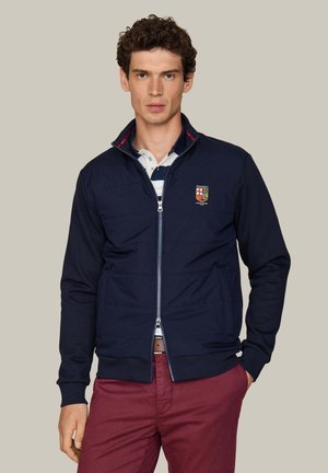 Jonge man met krullend haar draagt een marineblauw jack met rits, een wit en marineblauw gestreepte polo en bordeauxrode broek, staand tegen een effen achtergrond.