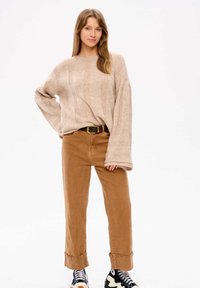 Beige gestrickter Pullover mit Rippstruktur, kombiniert mit braunen, hoch taillierten Hosen. Die Schuhe haben ein gewelltes Muster in Schwarz und Creme.
