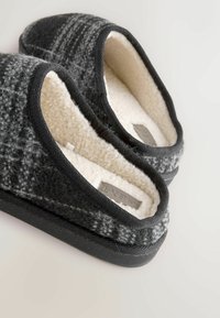 Mustat ja harmaat ruudulliset tossut, jotka on valmistettu villasta ja joissa on pehmeä beige fleece-vuori sekä kumipohja. Reunat on viimeistelty mustalla trimmillä.